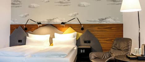A hotel room with a bed, bedside lamps, a chair, a table, and a wall with a cloud pattern.