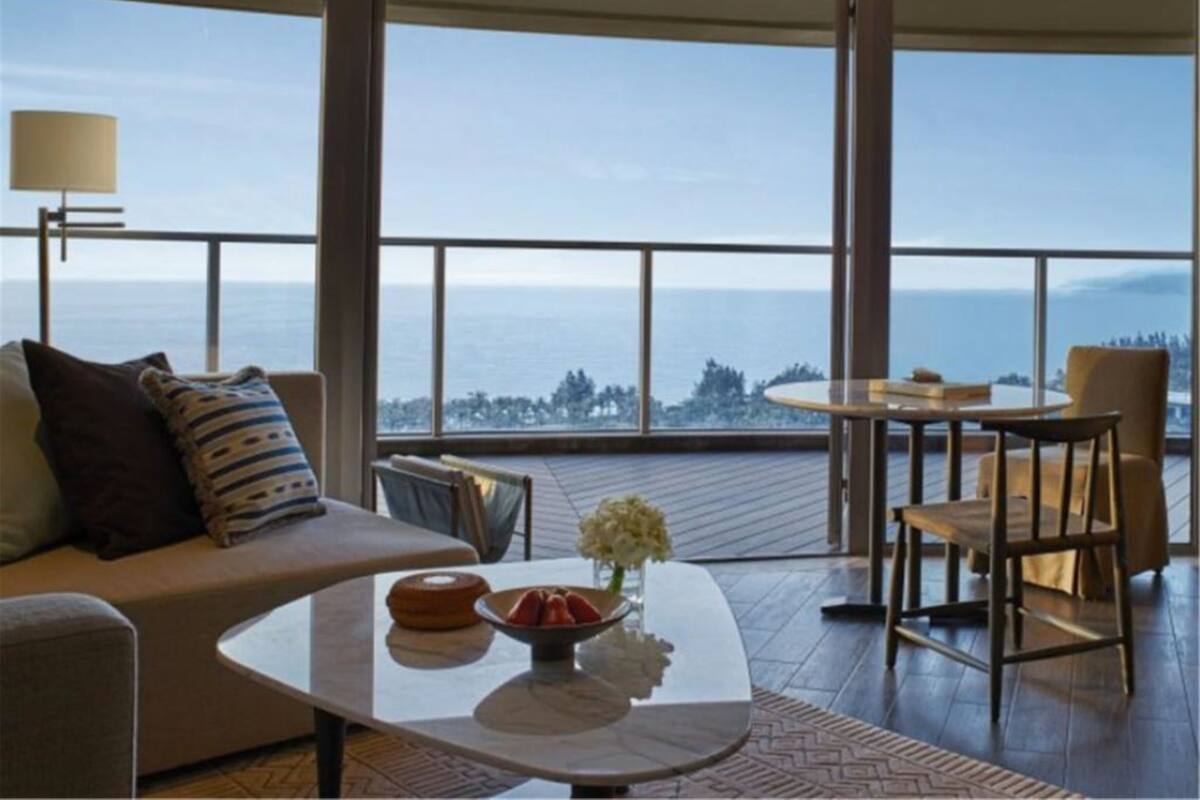 Una sala de estar moderna con un sofá, una mesa de centro y sillas, que cuenta con un amplio ventanal con vistas al mar.