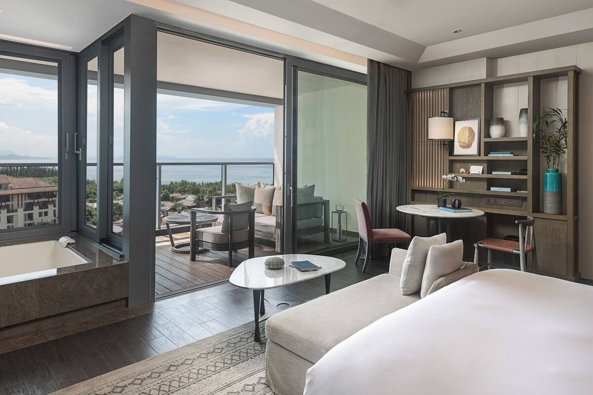 Una habitación de hotel moderna con una cama grande, una zona de estar con una mesa, una pequeña cocina y un balcón con vistas a la ciudad.