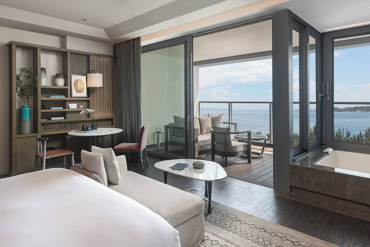 Una habitación de hotel moderna con una cama grande, un escritorio, una mesa redonda y un balcón con vistas al mar.