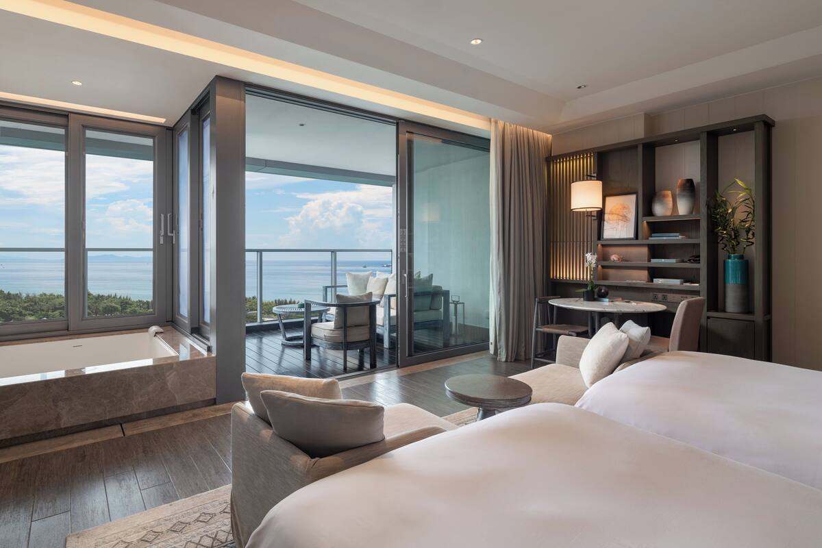 Una habitación de hotel moderna con una cama grande, una zona de estar con una mesa y un balcón con vistas al mar.