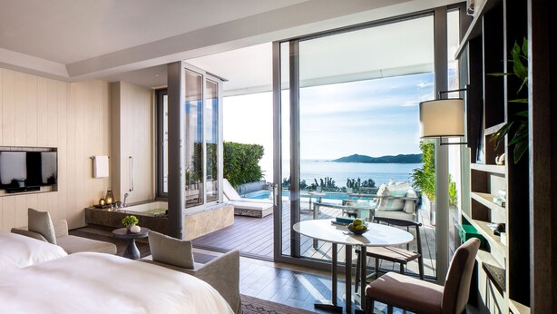 Una habitación de hotel moderna con una cama grande, un televisor, un balcón con vistas al mar y una mesita con una lámpara.