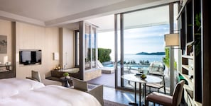 Una habitación de hotel moderna con una cama grande, un televisor, un balcón con vistas al mar y una mesita con una lámpara.