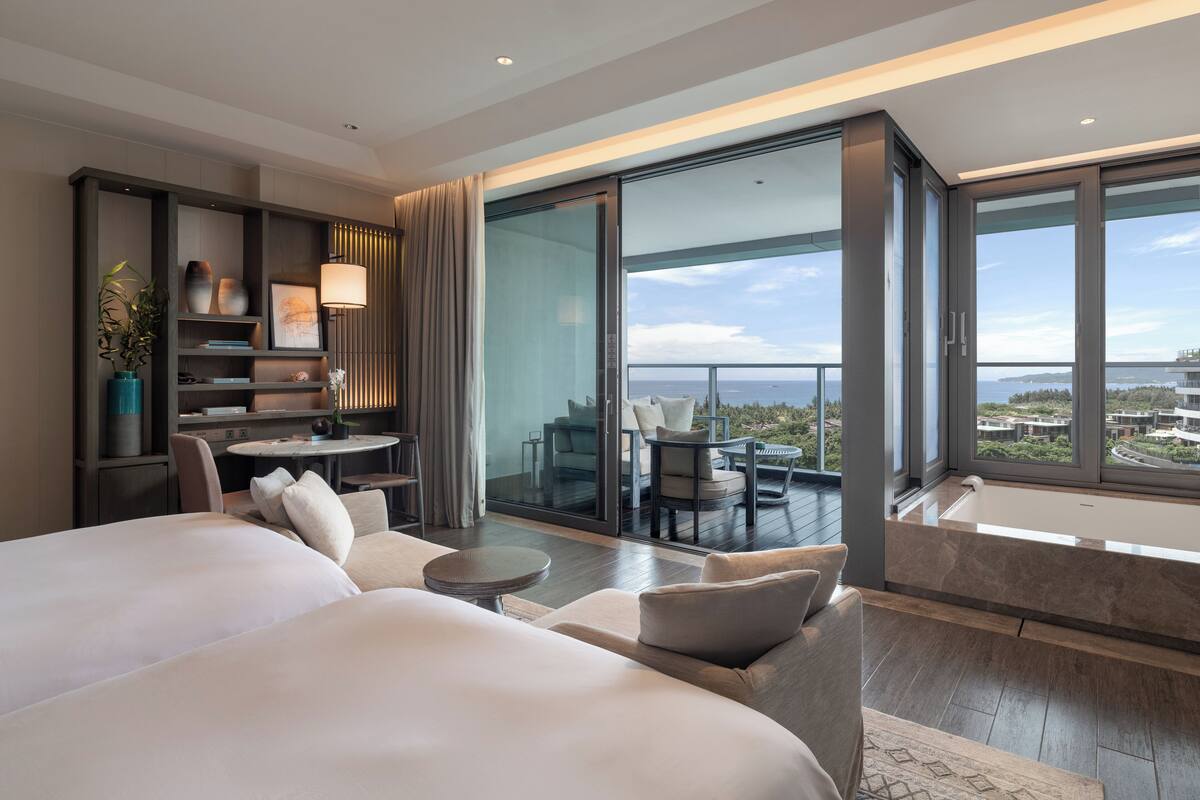 Una habitación de hotel moderna con una cama grande, una zona de estar con una mesa redonda, una pequeña estantería y un balcón con vistas a la ciudad.