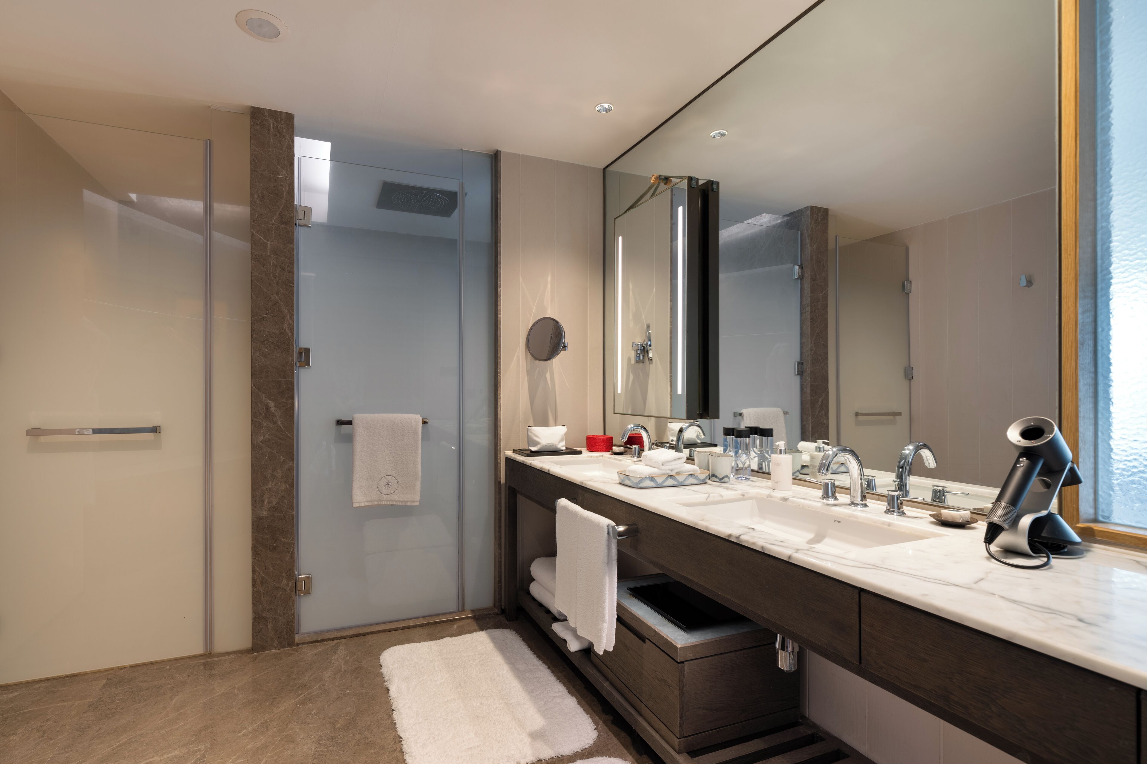 A modern bathroom with a glass shower enclosure, a large mirror, dual sinks, and a towel rack.