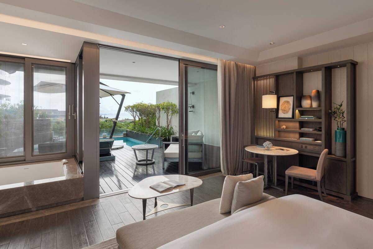 Una habitación de hotel moderna con una cama grande, una pequeña mesa redonda, un sofá, un escritorio con una silla y un vidrio que da a un espacio exterior con piscina y sillas de descanso.