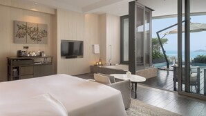 Una habitación de hotel moderna con una cama grande, un escritorio con televisor de pantalla plana, un pequeño rincón de descanso y un balcón con vistas al mar.