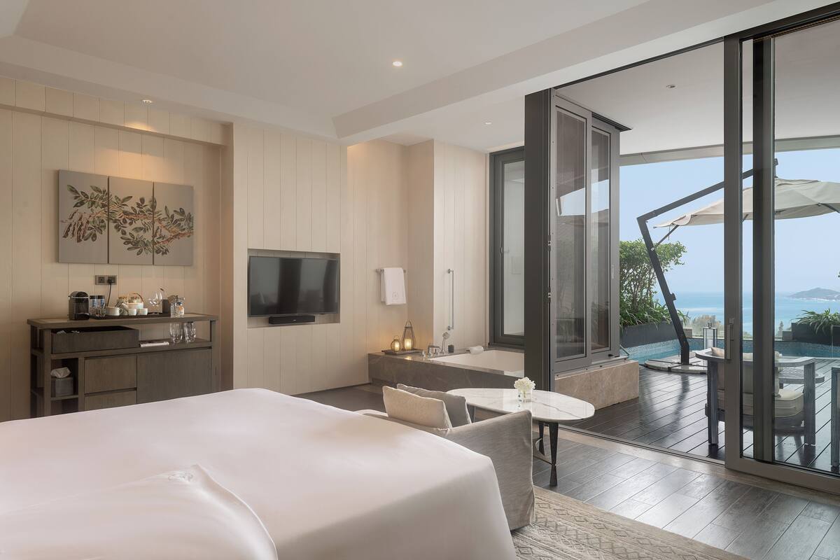 Una habitación de hotel moderna con una cama grande, un escritorio con televisor de pantalla plana, un pequeño rincón de descanso y un balcón con vistas al mar.