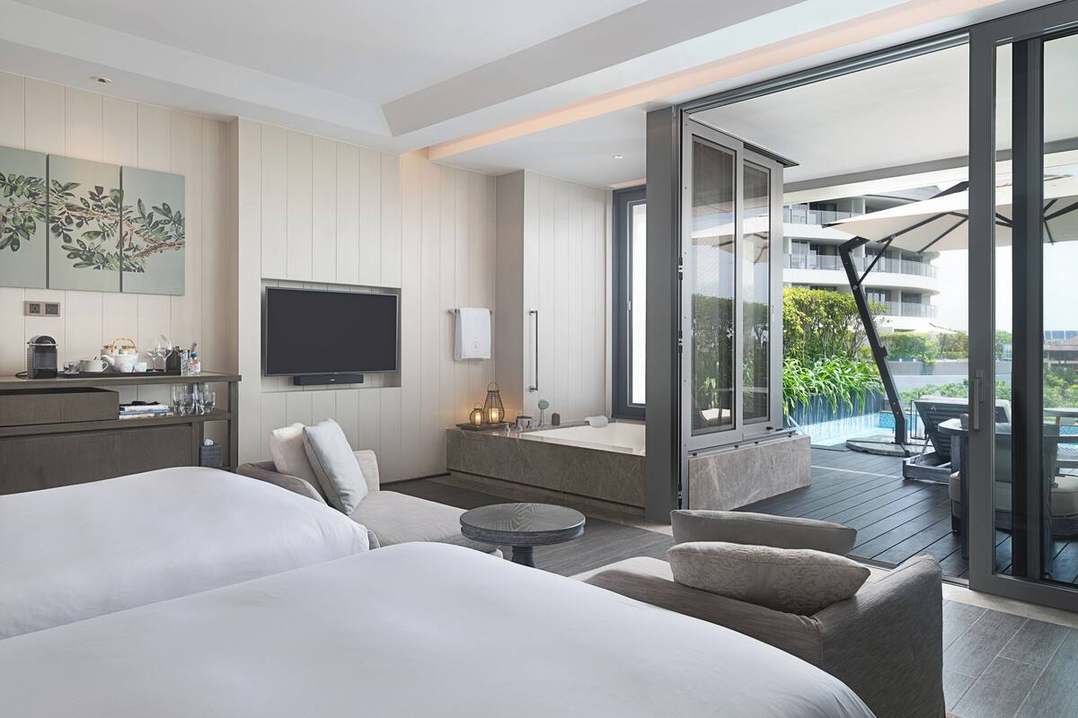 Una habitación de hotel moderna con una cama grande, un sofá, una mesita redonda, un televisor y un balcón con mobiliario exterior.