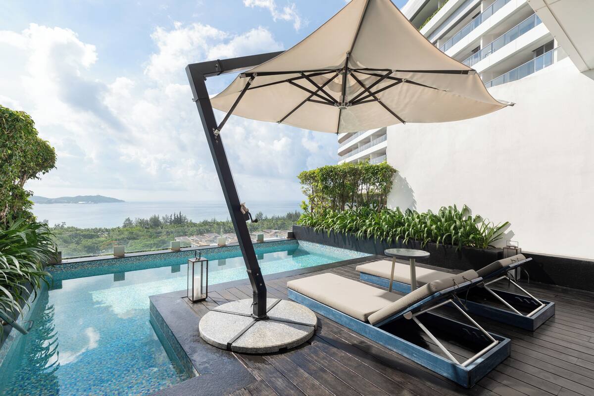 Piscina infinita en la azotea con sillas de descanso y un toldo, con vistas a un paisaje urbano y al mar.