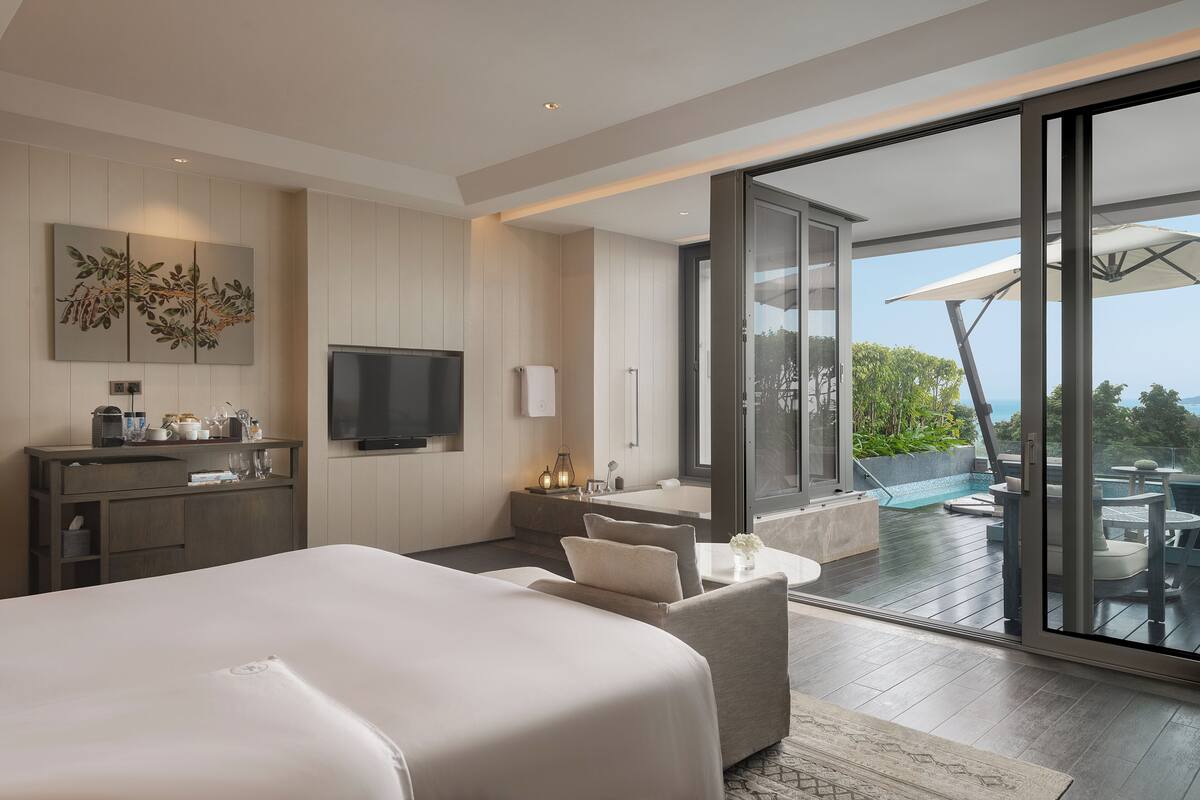 Una habitación de hotel moderna con una cama grande, televisión, baño con bañera y balcón con mobiliario exterior.