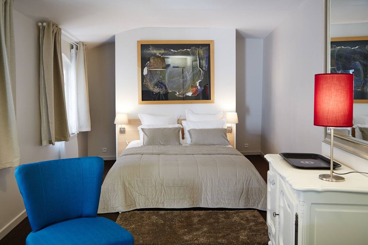 A hotel room with a bed, a blue chair, a red lamp, and a painting on the wall.