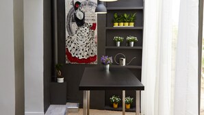 Un interior moderno con una mesa de madera oscura, dos lámparas colgantes negras, una cortina blanca y una pared con una obra de arte grande enmarcada y estantes con plantas en macetas.