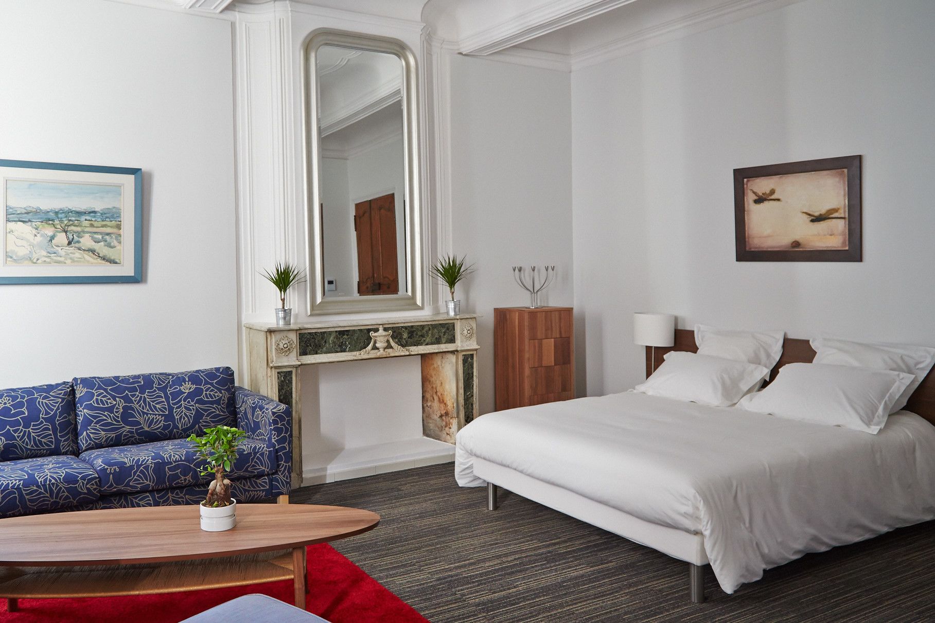 A hotel room with a bed, a blue sofa, a small table with a plant, a fireplace, and a painting on the wall.