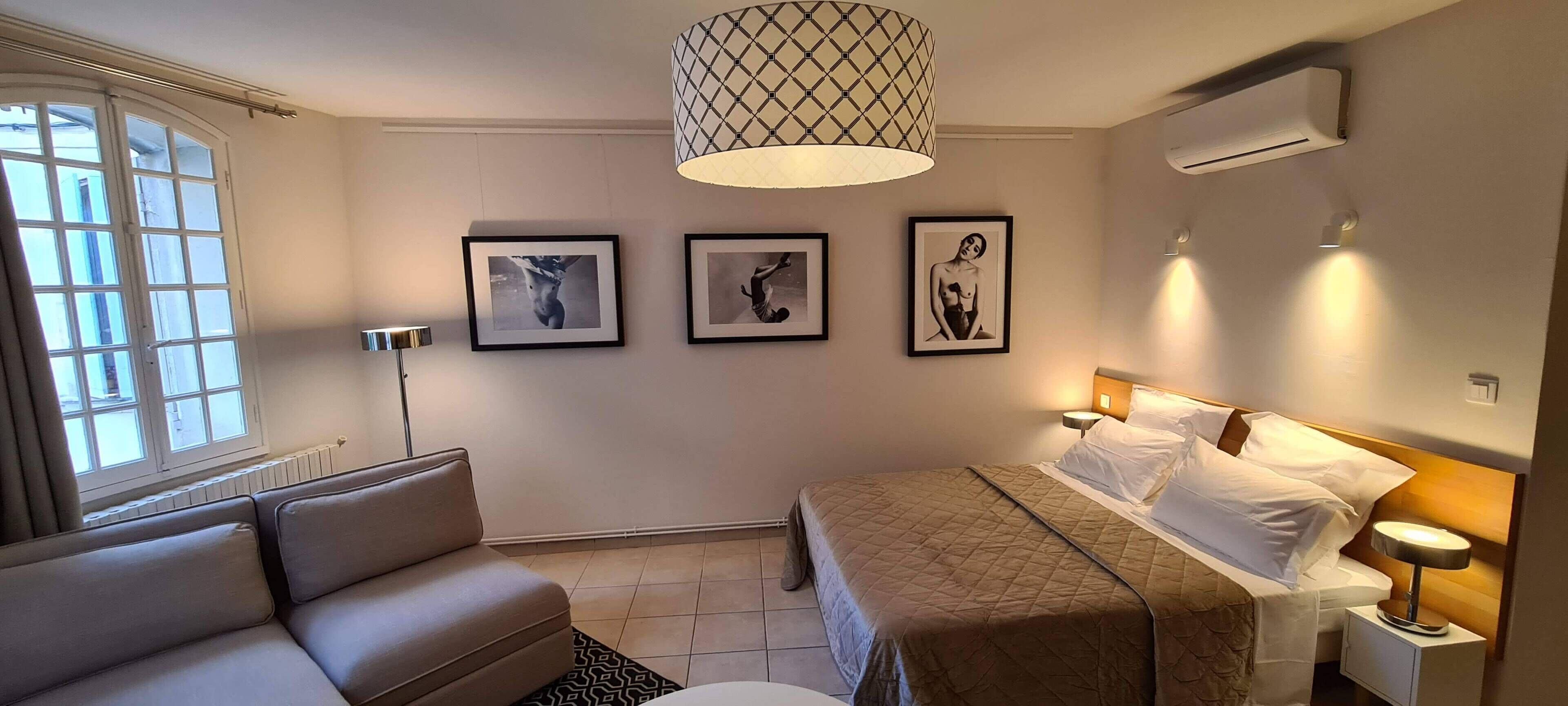 A hotel room with a bed, a sofa, a floor lamp, and three framed pictures on the wall.