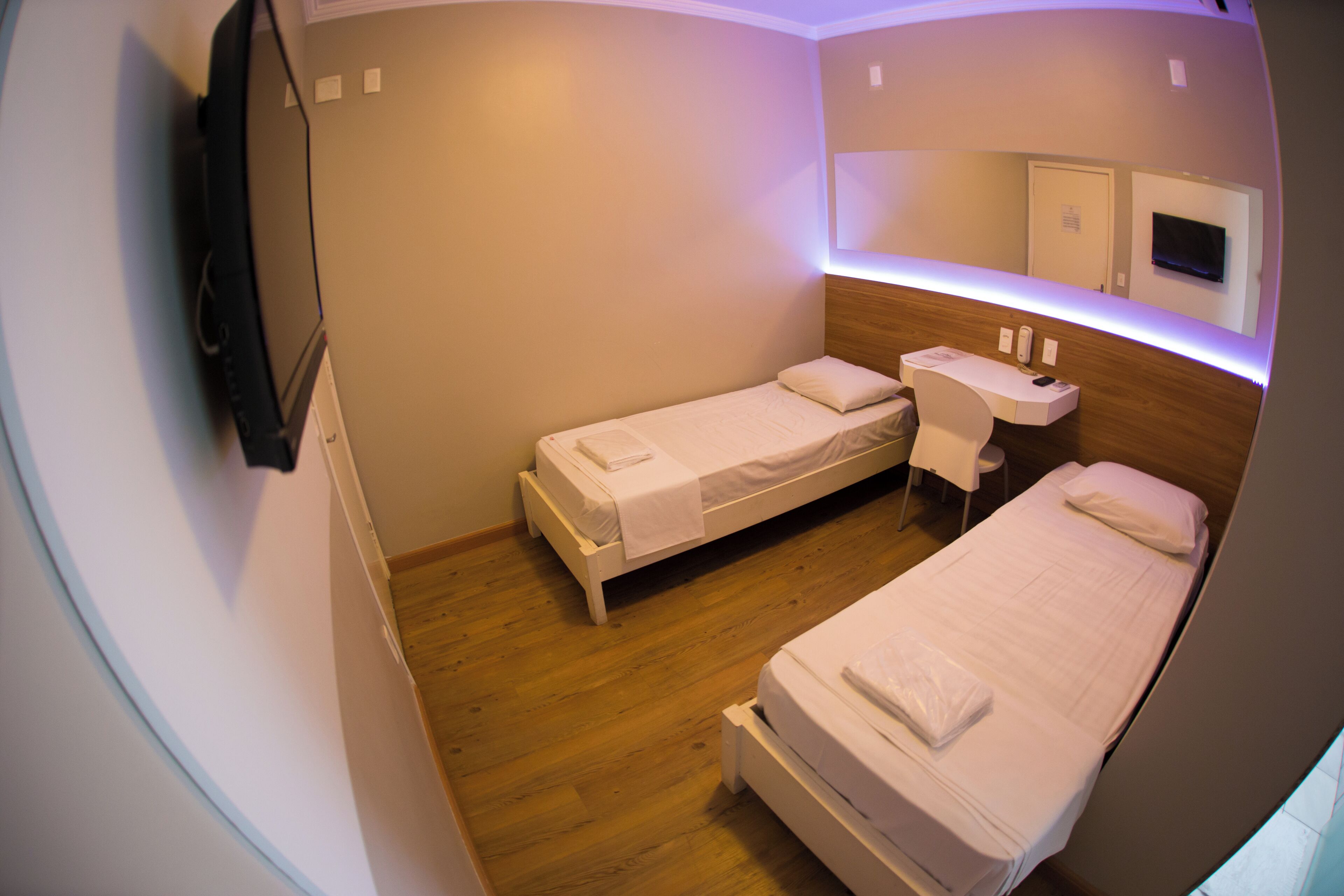 Une chambre d’hôtel de taille réduite, équipée de deux lits simples, d’un petit bureau, d’une chaise, d’un miroir et d’un téléviseur fixé au mur.