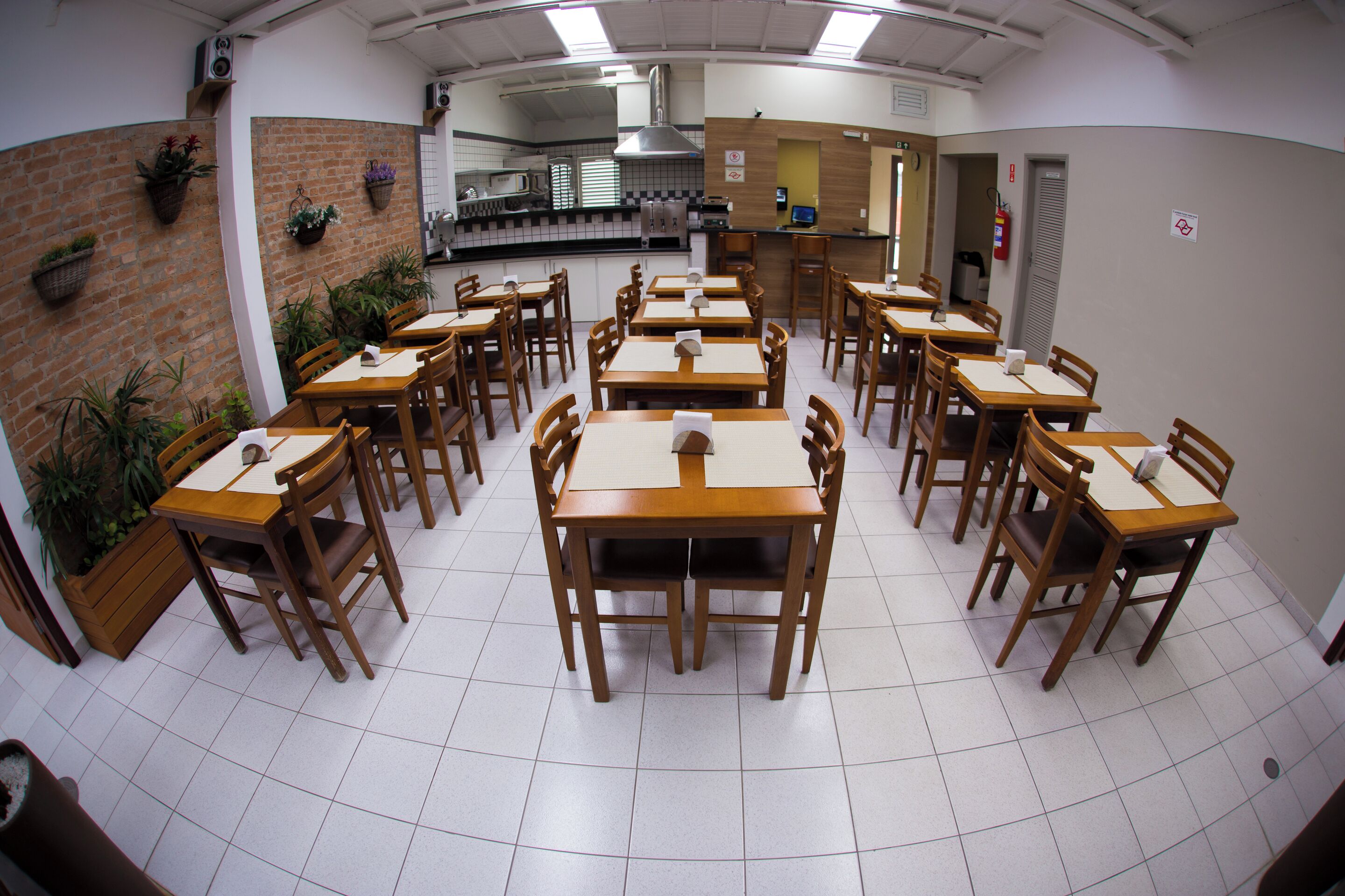 Un espace repas avec plusieurs tables et chaises en bois, une cuisine à l’arrière-plan et des plantes en pot disposées sur les murs.