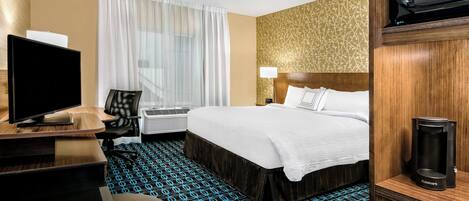 A hotel room with a bed, a desk with a computer, a television, a coffee maker, and a patterned carpet.