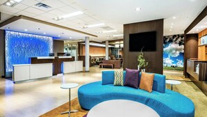 Un hall d’hôtel moderne avec un canapé bleu, un comptoir d’accueil, un téléviseur à écran plat et une fresque représentant un ciel bleu avec des nuages.