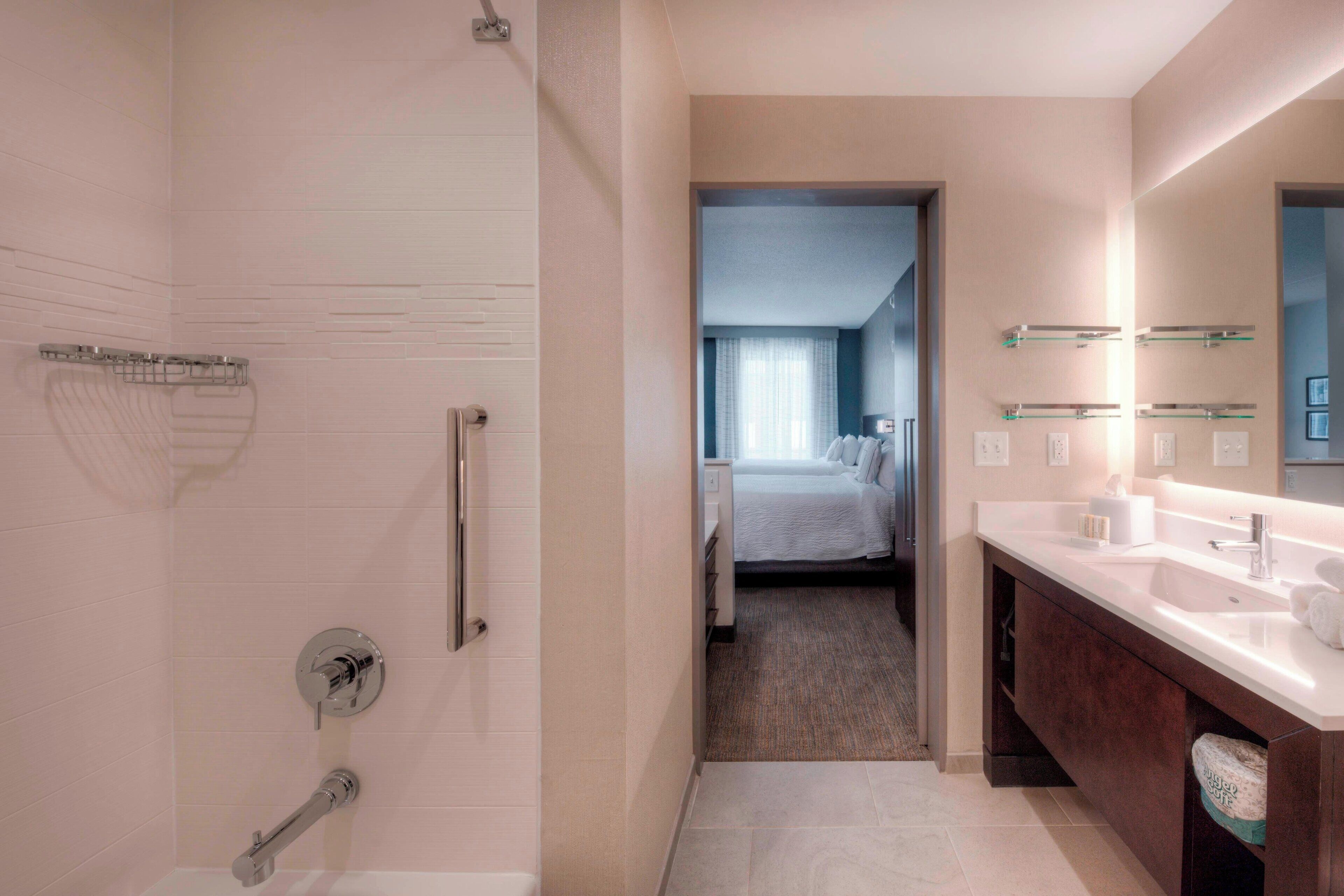 A modern bathroom with a shower, toilet, and vanity. There is a bed visible through an open door in the background.