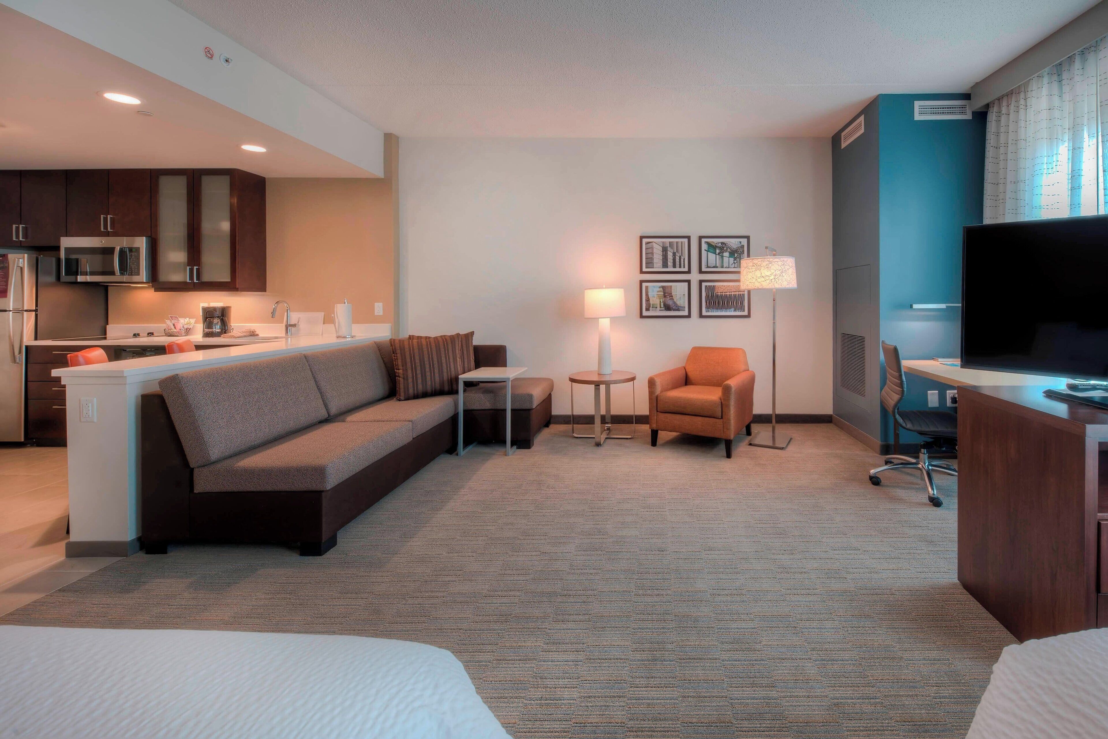 A hotel room with a kitchenette, a sofa, a chair, a small table, a lamp, a TV, and a bed.