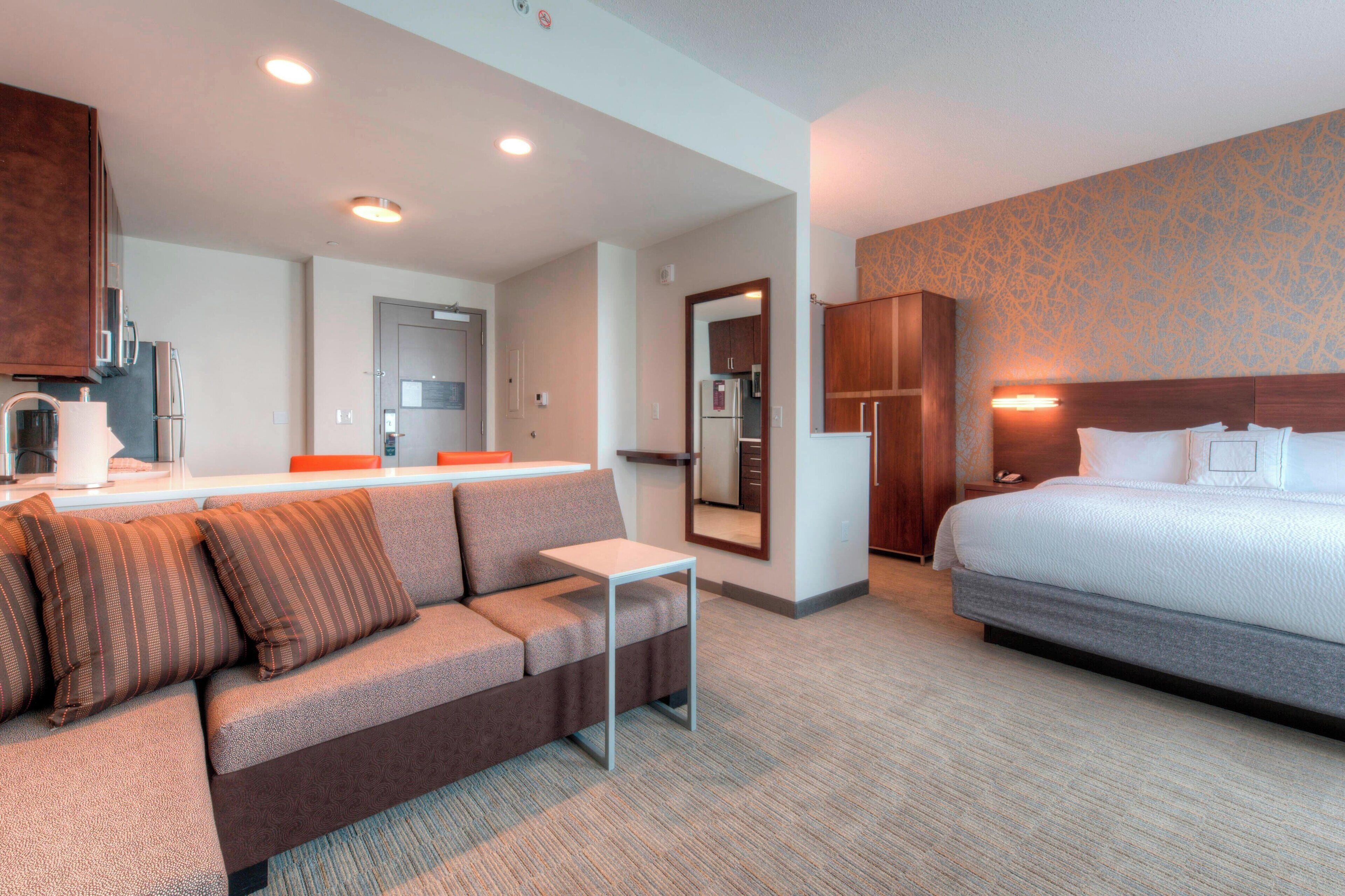 A hotel room with a sofa, a bed, a small table, a mirror, and a kitchenette.