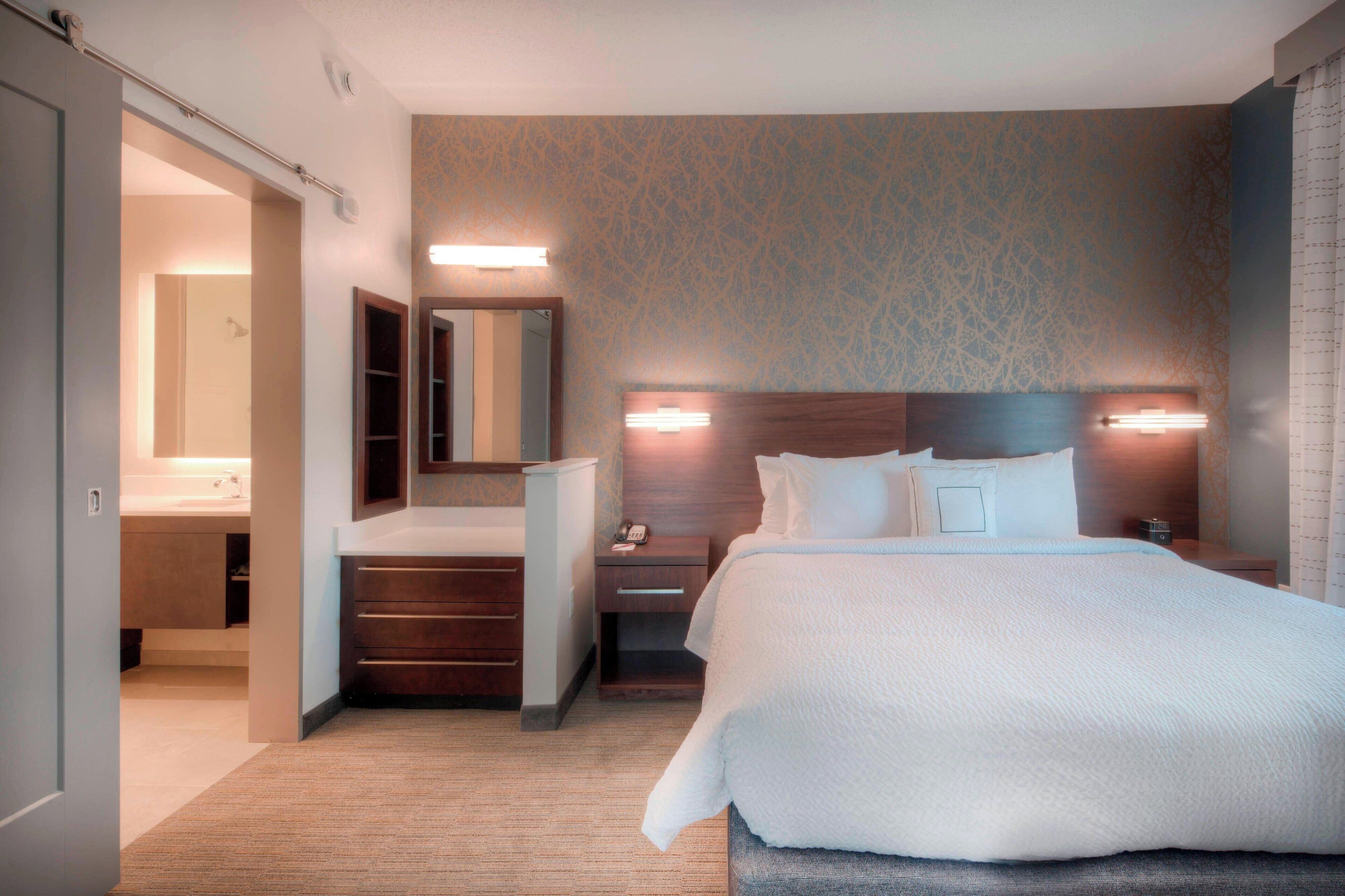 Une chambre d’hôtel avec un grand lit, une tête de lit en bois, une table de chevet, une commode, un miroir, une armoire et une salle de bain visible à travers une porte ouverte.