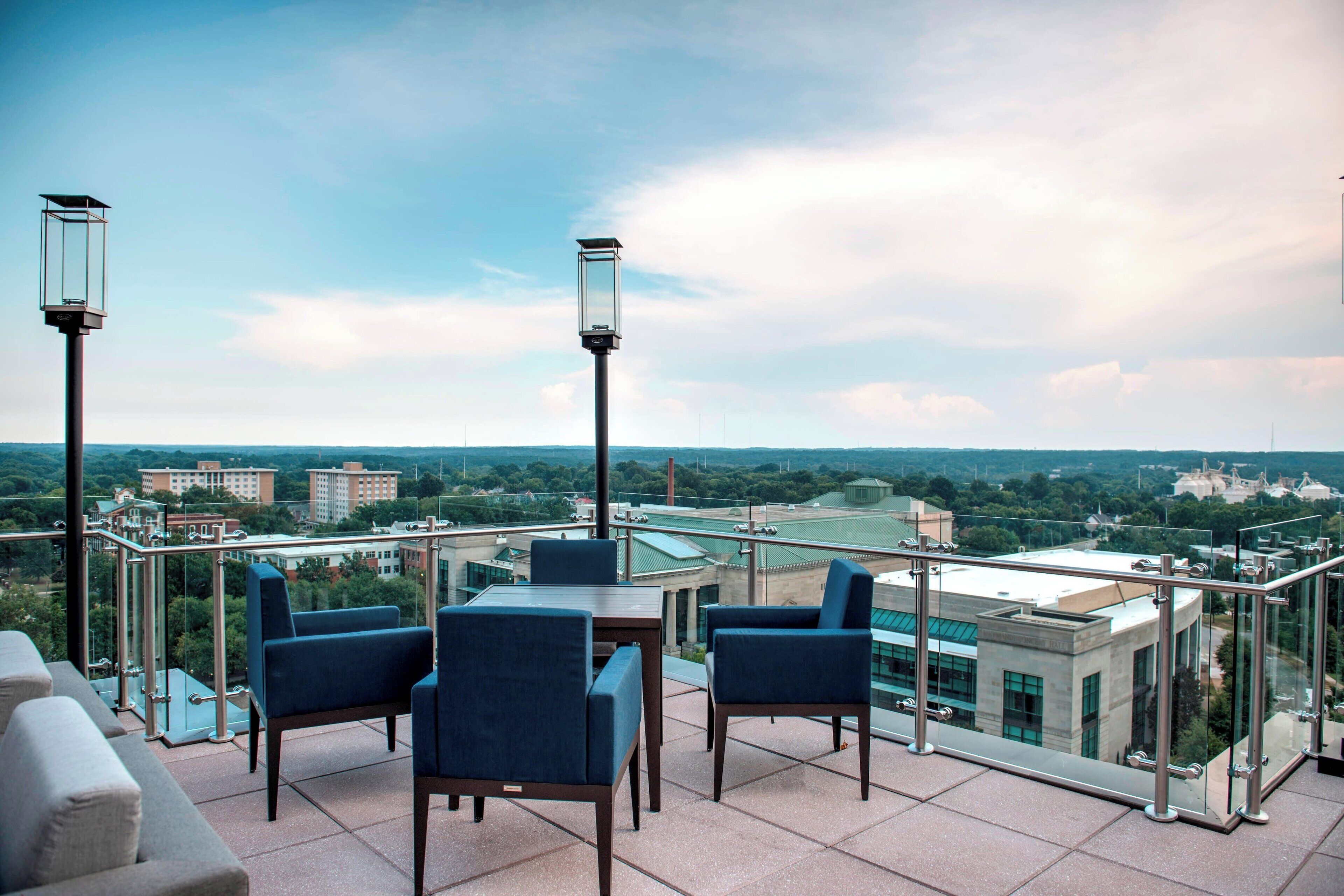 A rooftop terrace with a glass balustrade, a table with chairs, and two street lamps.