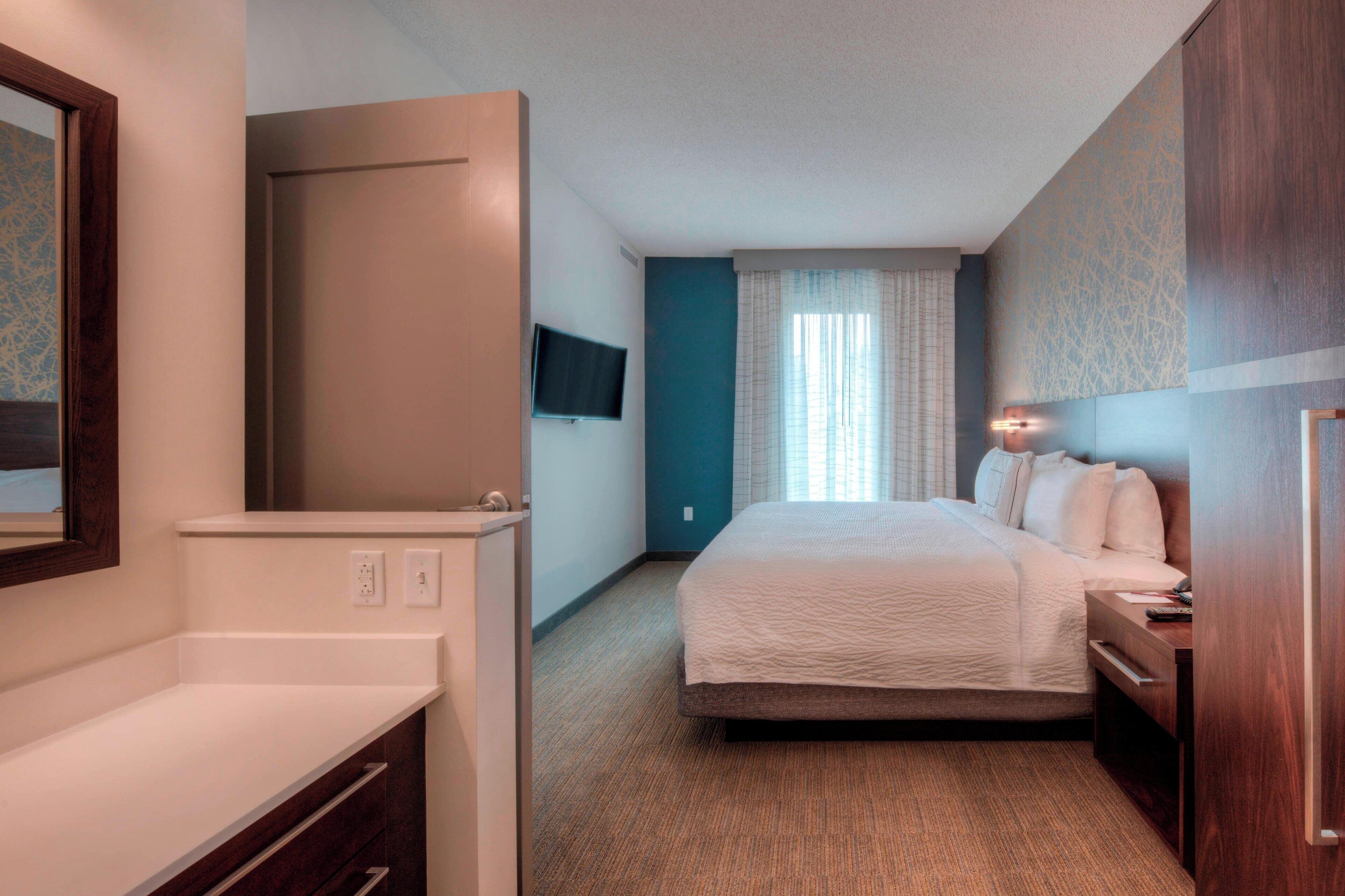 A hotel room with a bed, a desk, a TV mounted on the wall, and a window with curtains.