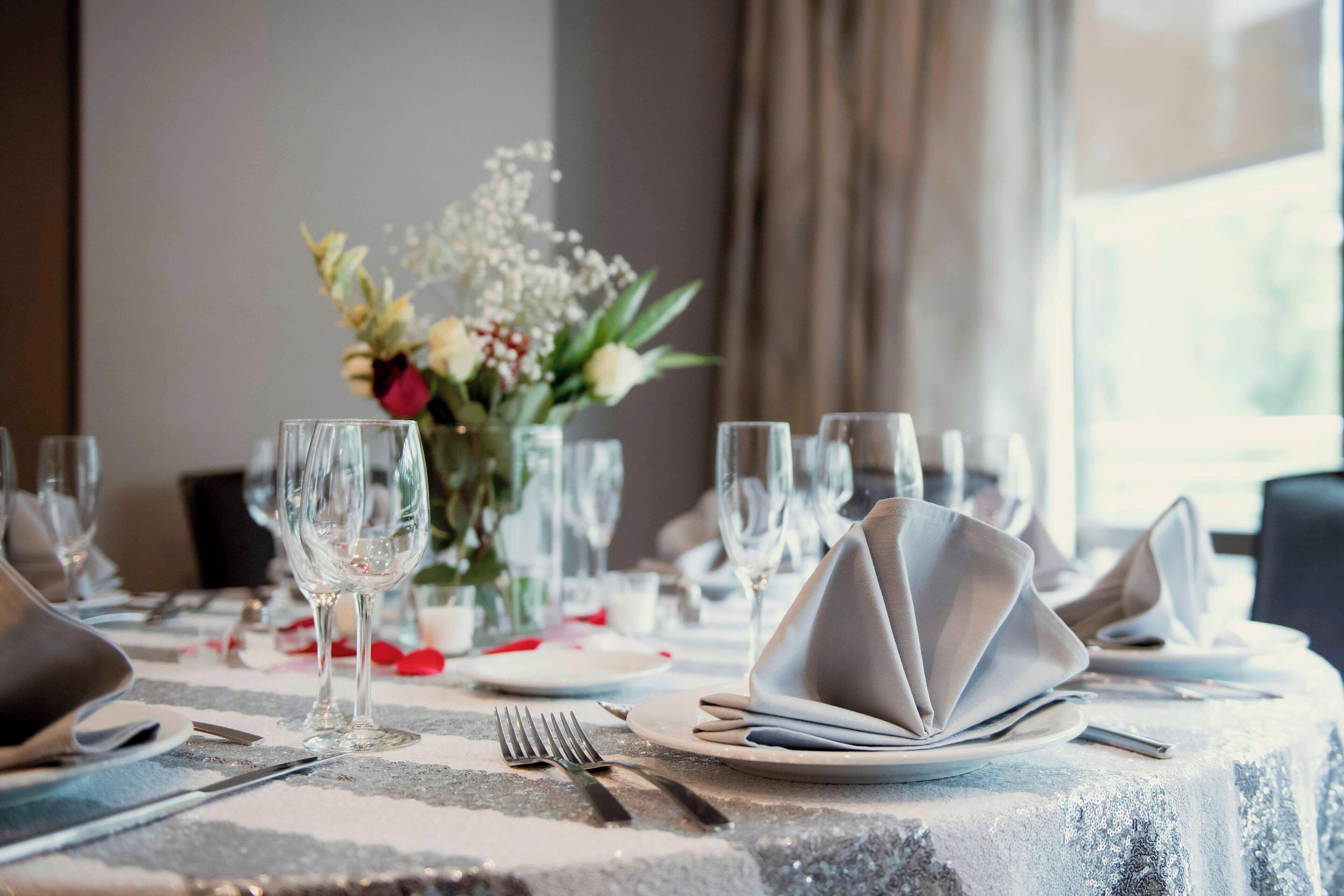 Una mesa de comedor dispuesta con servilletas blancas, cubiertos y copas de vino, con un centro de mesa decorativo.