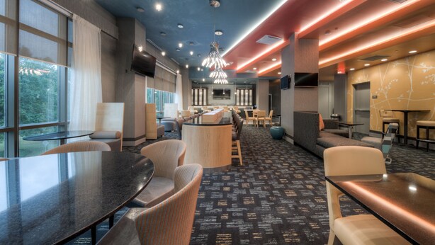 Un hall d’hôtel moderne doté d’un bar central, de différents espaces aménagés pour s’asseoir et d’un grand écran de télévision.