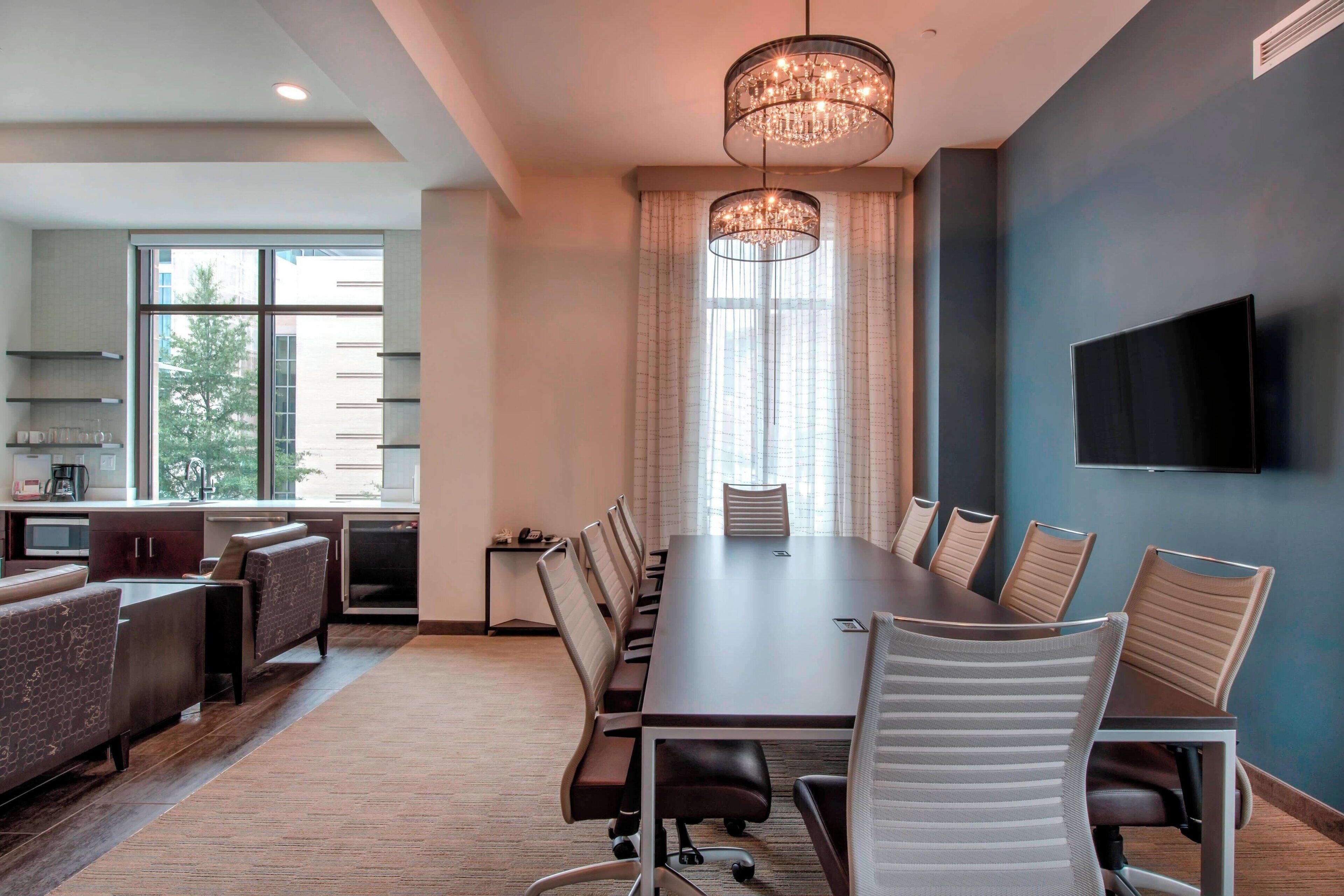 A modern meeting room with a long table, comfortable chairs, a chandelier, and a television on the wall.