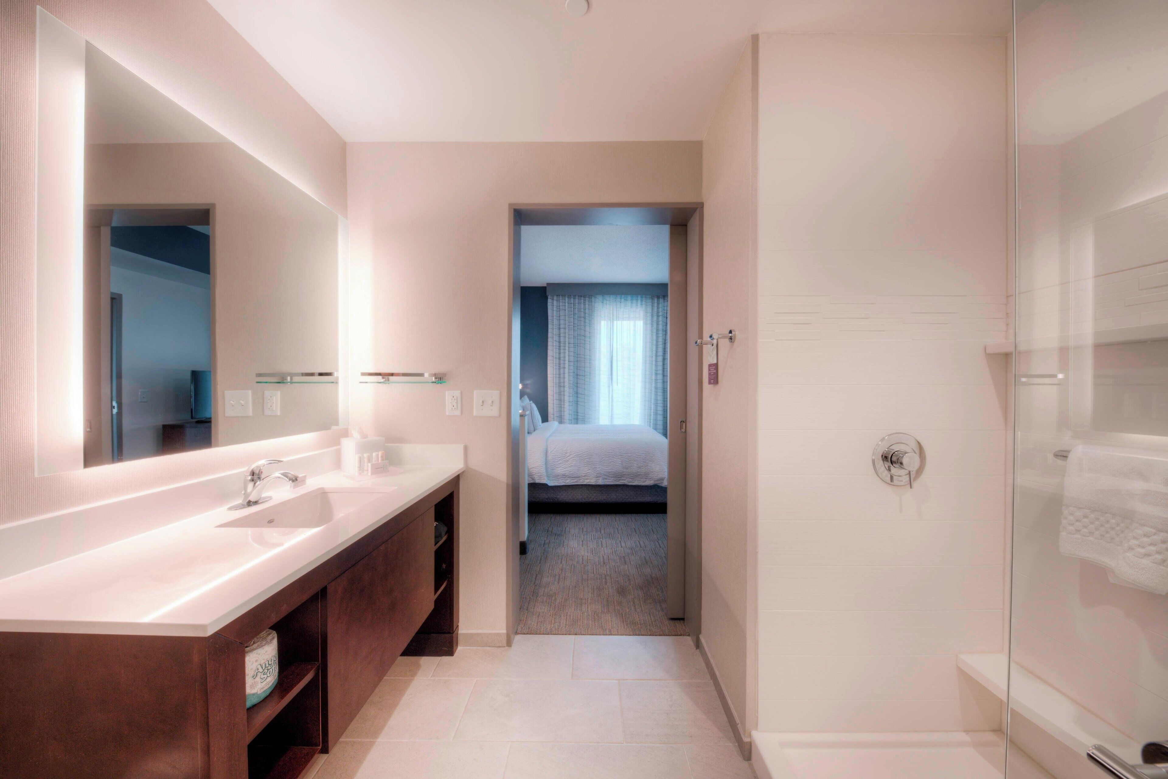 Un baño moderno con un espejo grande, dos lavamanos, ducha y una habitación visible a través de una puerta abierta.