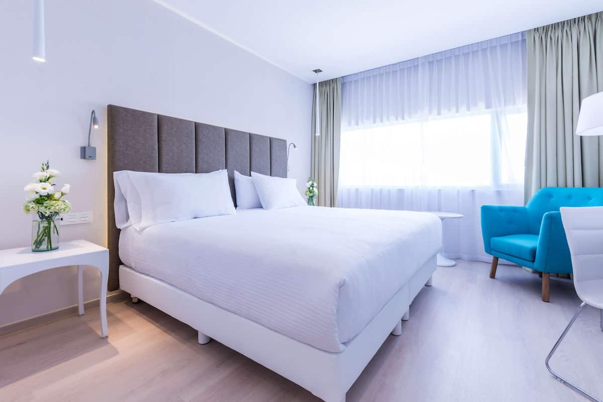 A modern hotel room with a large bed, a small side table, a blue armchair, and a white desk.