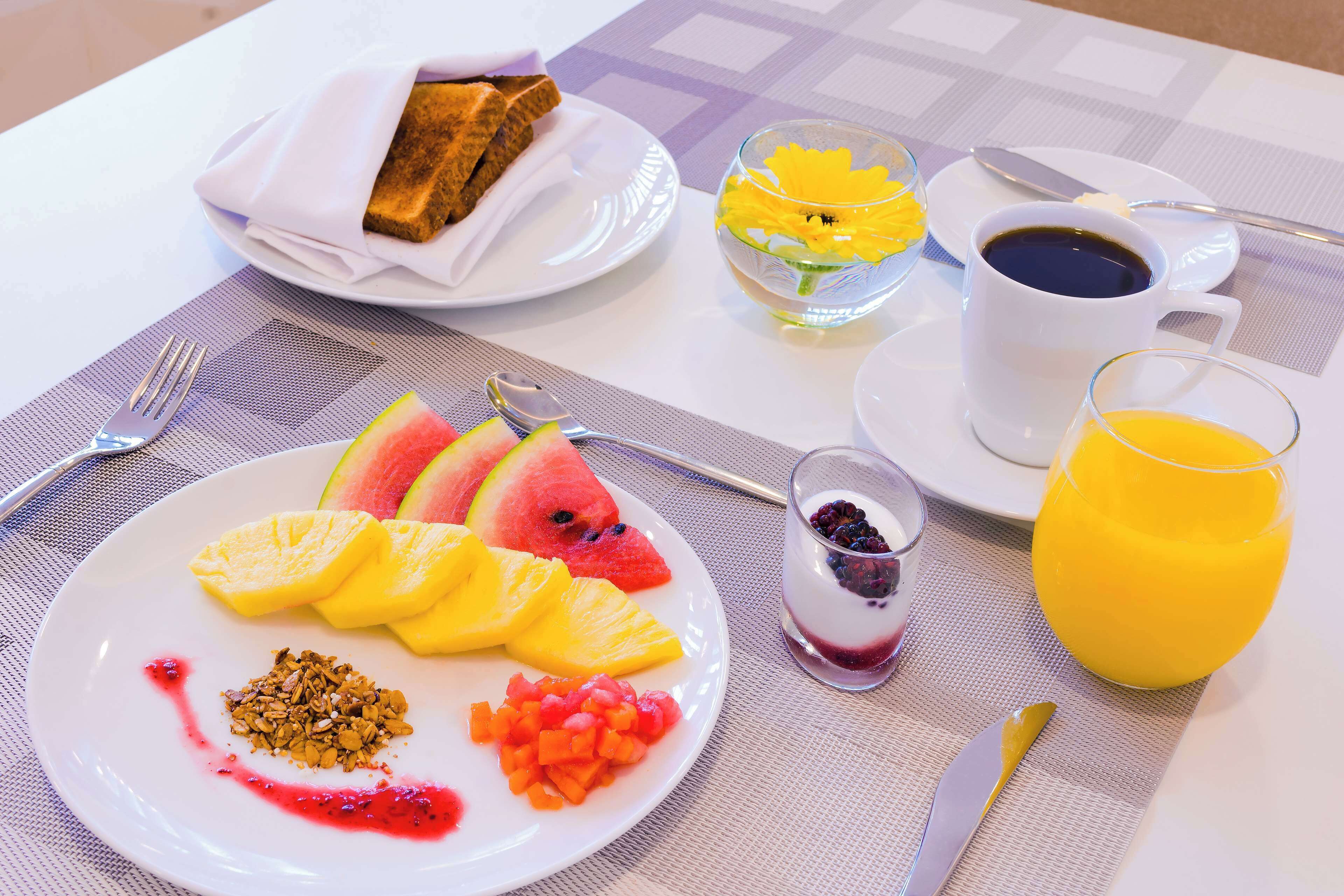 Un desayuno con un plato de fruta, un vaso de zumo de naranja, una taza de café y un bol pequeño de yogur con frutos rojos.