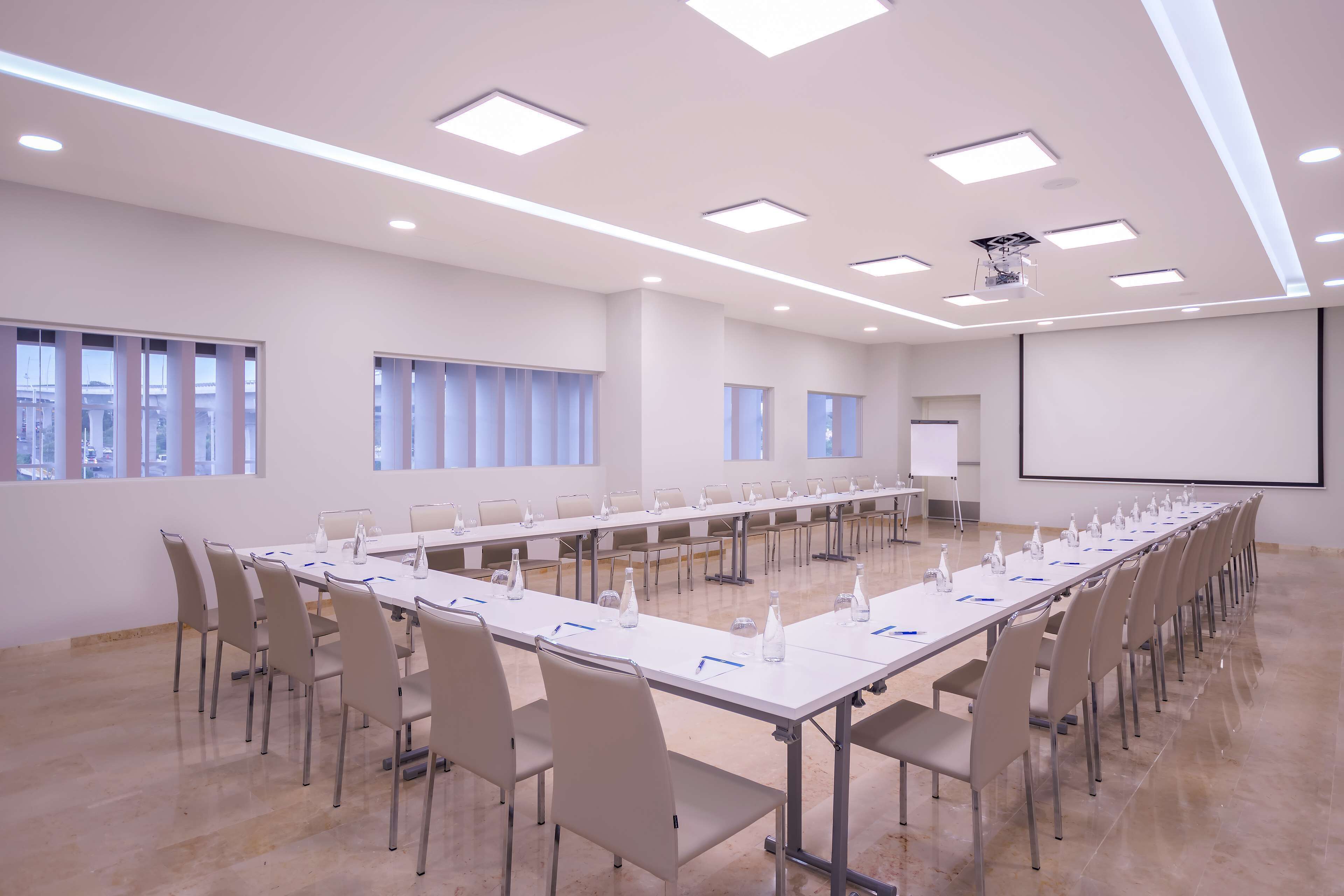 Ein Konferenzraum mit langem Tisch, Stühlen, einer Projektionsleinwand und Fenstern.