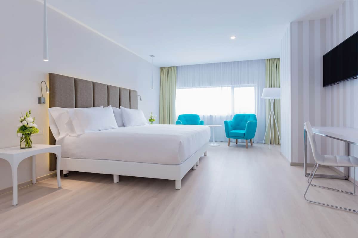 A modern hotel room with a large bed, a desk, two chairs, a TV, and a window with curtains.