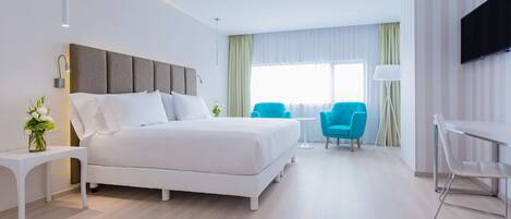 Habitación de hotel moderna con una cama grande, un escritorio, dos sillas, un televisor y una ventana con cortinas.