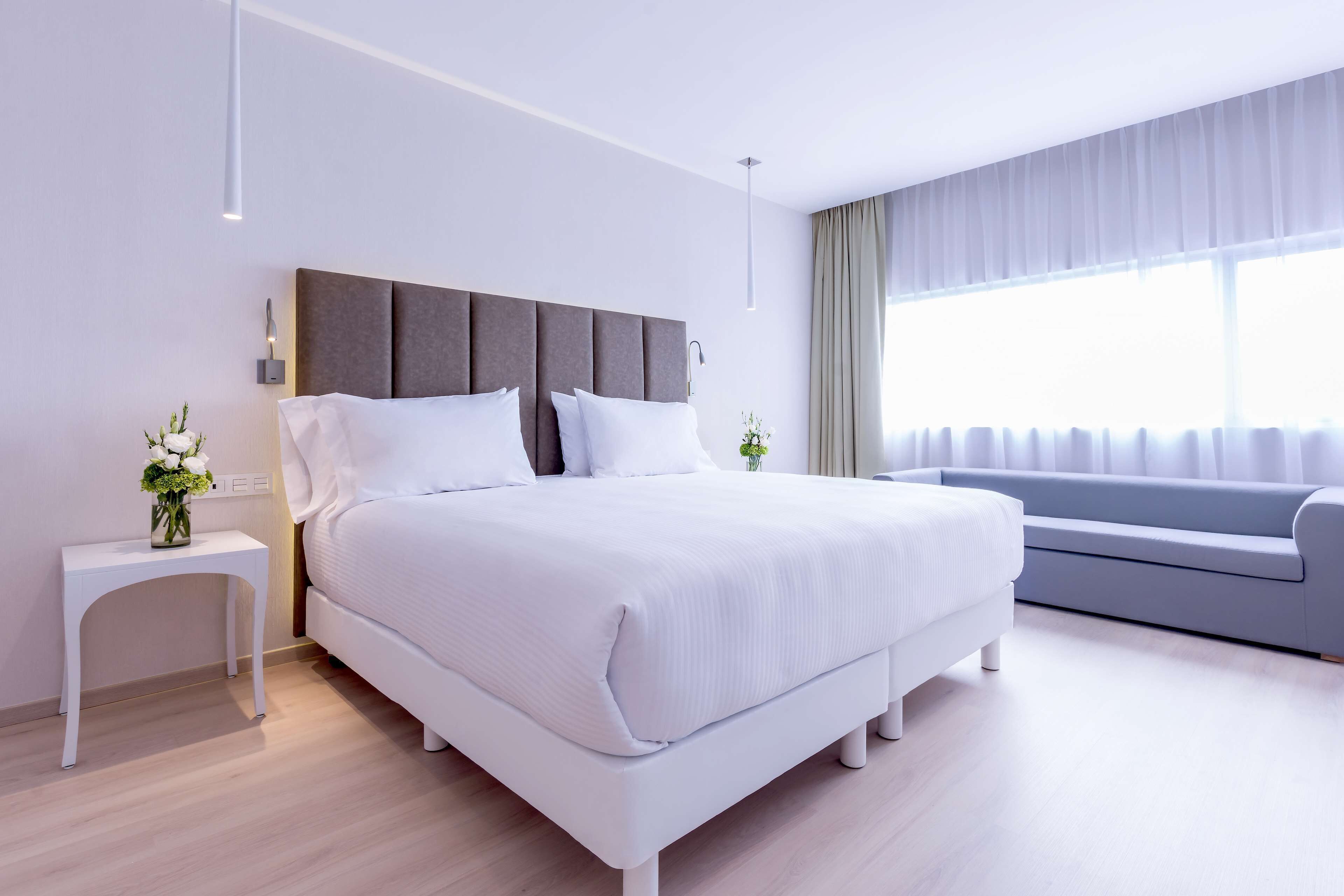 A modern bedroom with a large bed, a bedside table with a vase, a grey sofa, and a large window with curtains.
