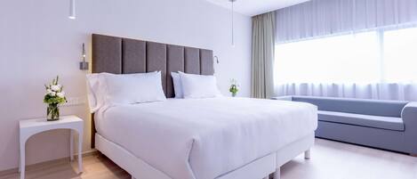 A modern bedroom with a large bed, a bedside table with a vase, a grey sofa, and a large window with curtains.