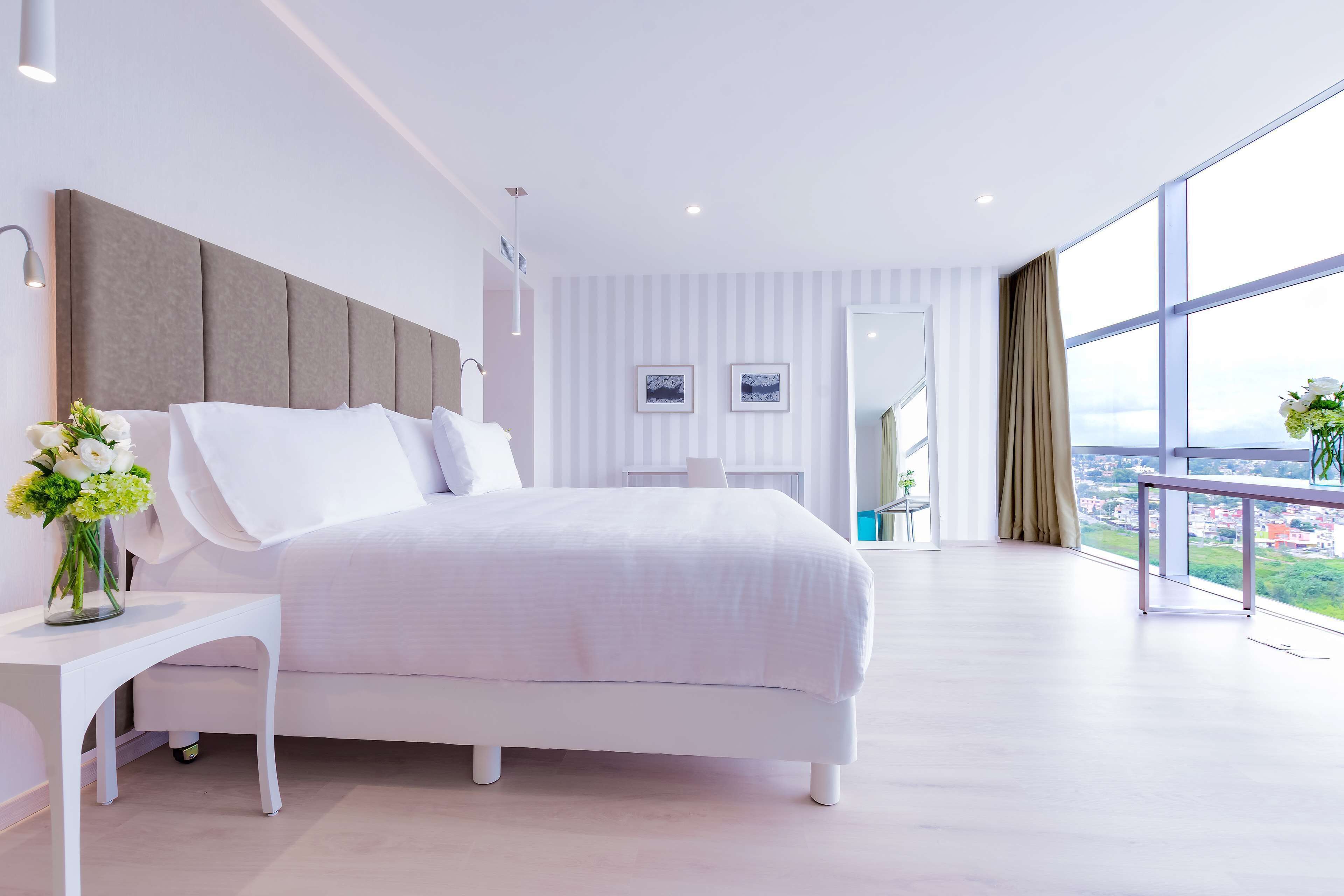 Een moderne slaapkamer met een groot bed, een nachtkastje met een bloemenvaas, en uitzicht op de stad door grote ramen.