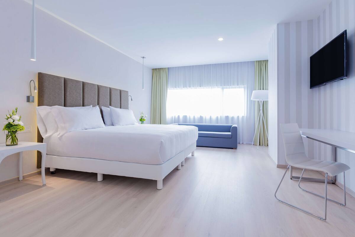 A modern hotel room with a large bed, a desk, a chair, a TV, and a window with curtains.