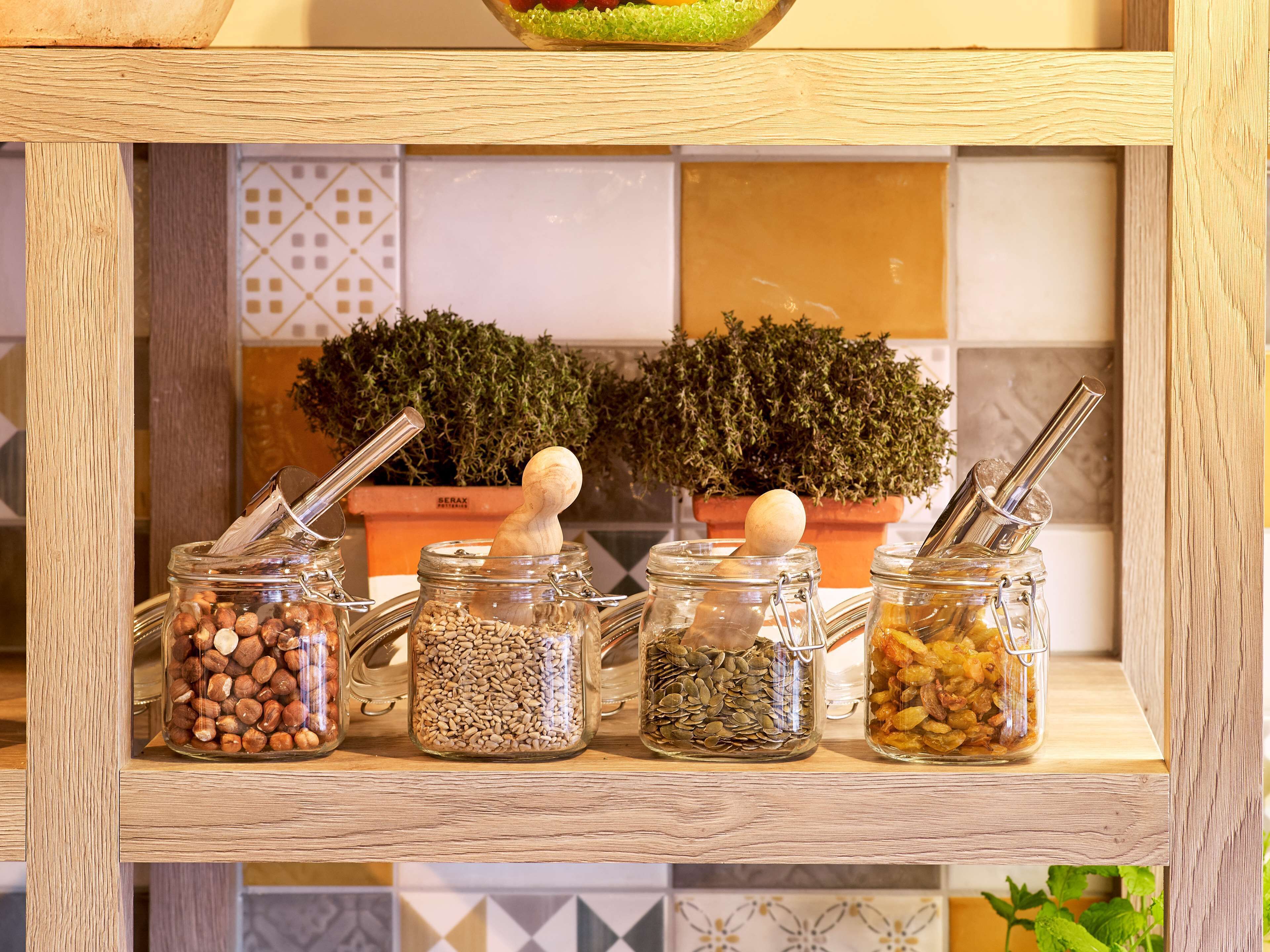 Una estantería de madera con frascos de vidrio que contienen diferentes nueces y semillas, junto a dos plantas en macetas.