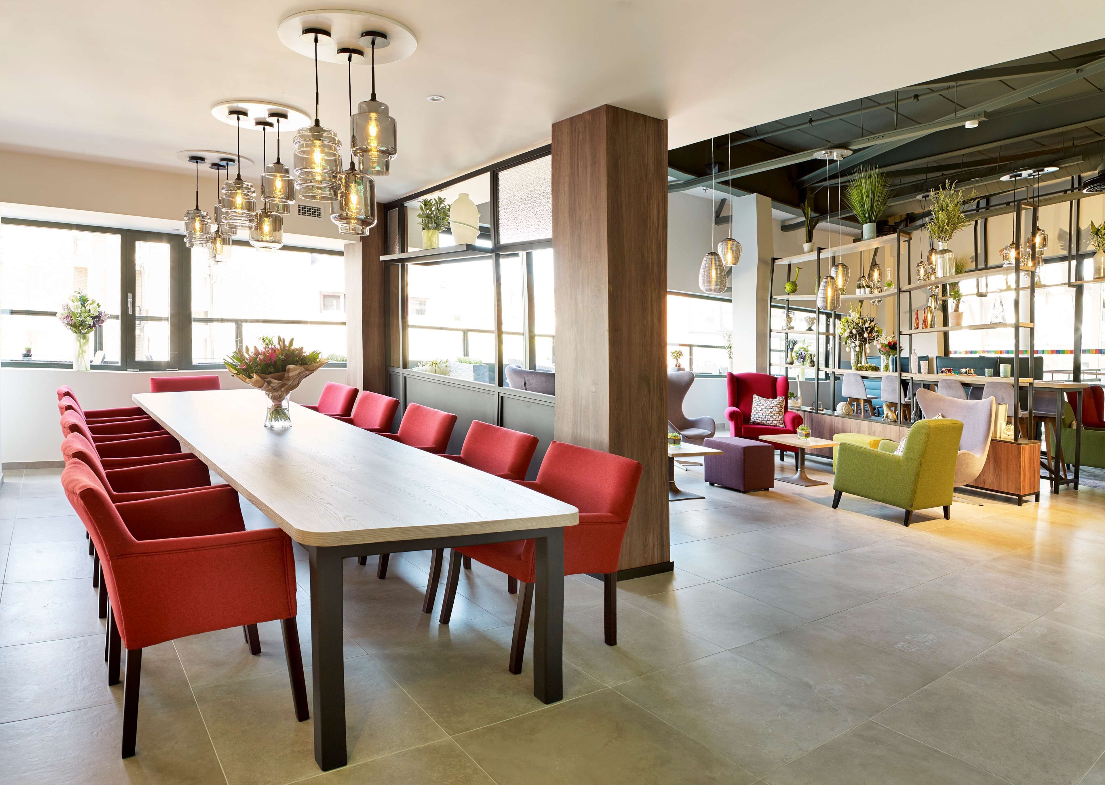 A modern dining area with a long table and red chairs, large windows, and hanging pendant lights.