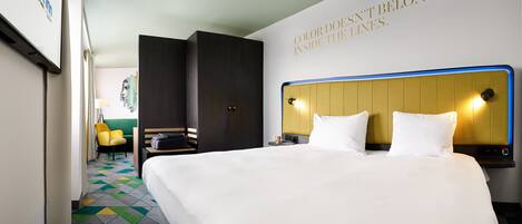 Ein Hotelzimmer mit einem großen Bett, einem Kopfteil mit integrierter Beleuchtung, einem Kleiderschrank, einer Sitzecke mit einem Sessel und einem kleinen Tisch sowie einer Wand mit einem motivierenden Zitat.
