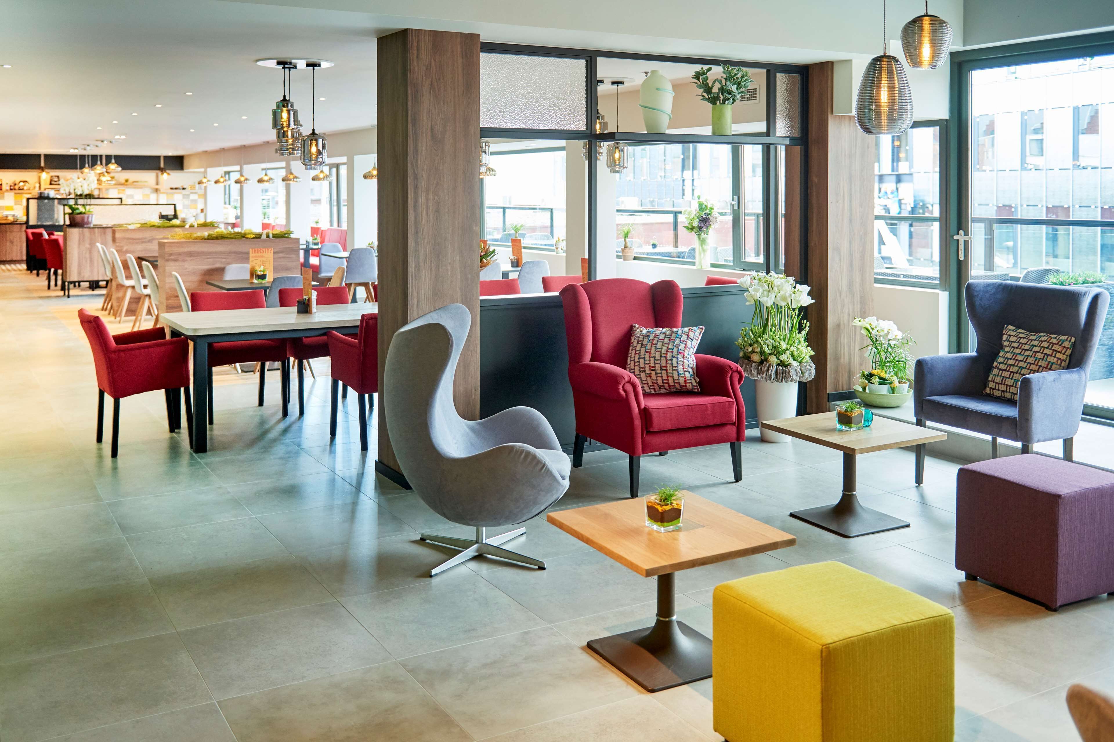 A modern hotel lobby with a mix of seating options, including a dining area with a long table and chairs, a variety of armchairs and sofas, and a bar area in the background.