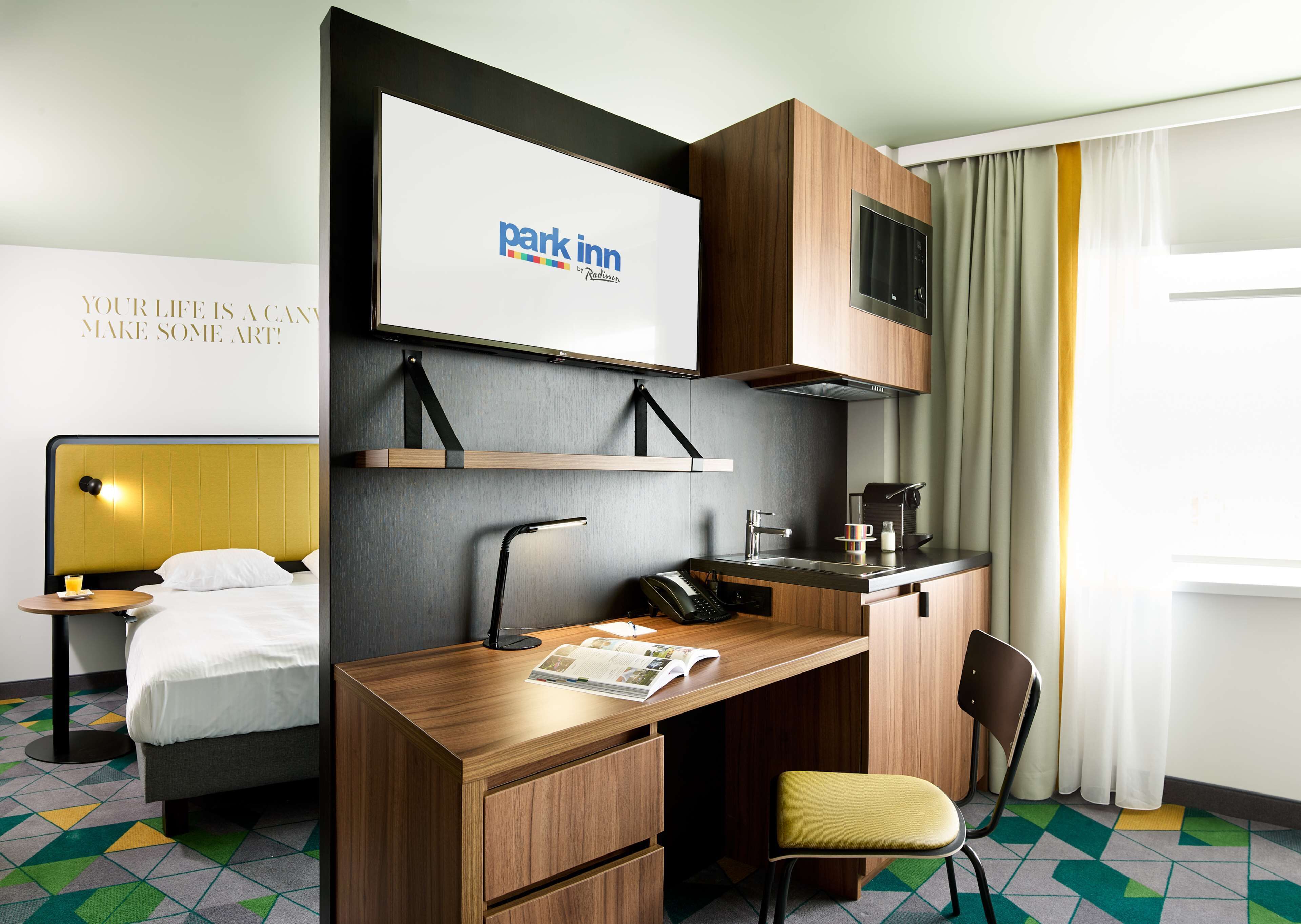 A hotel room with a bed, a desk with a chair, a small kitchenette, and a television displaying the Park Inn logo.