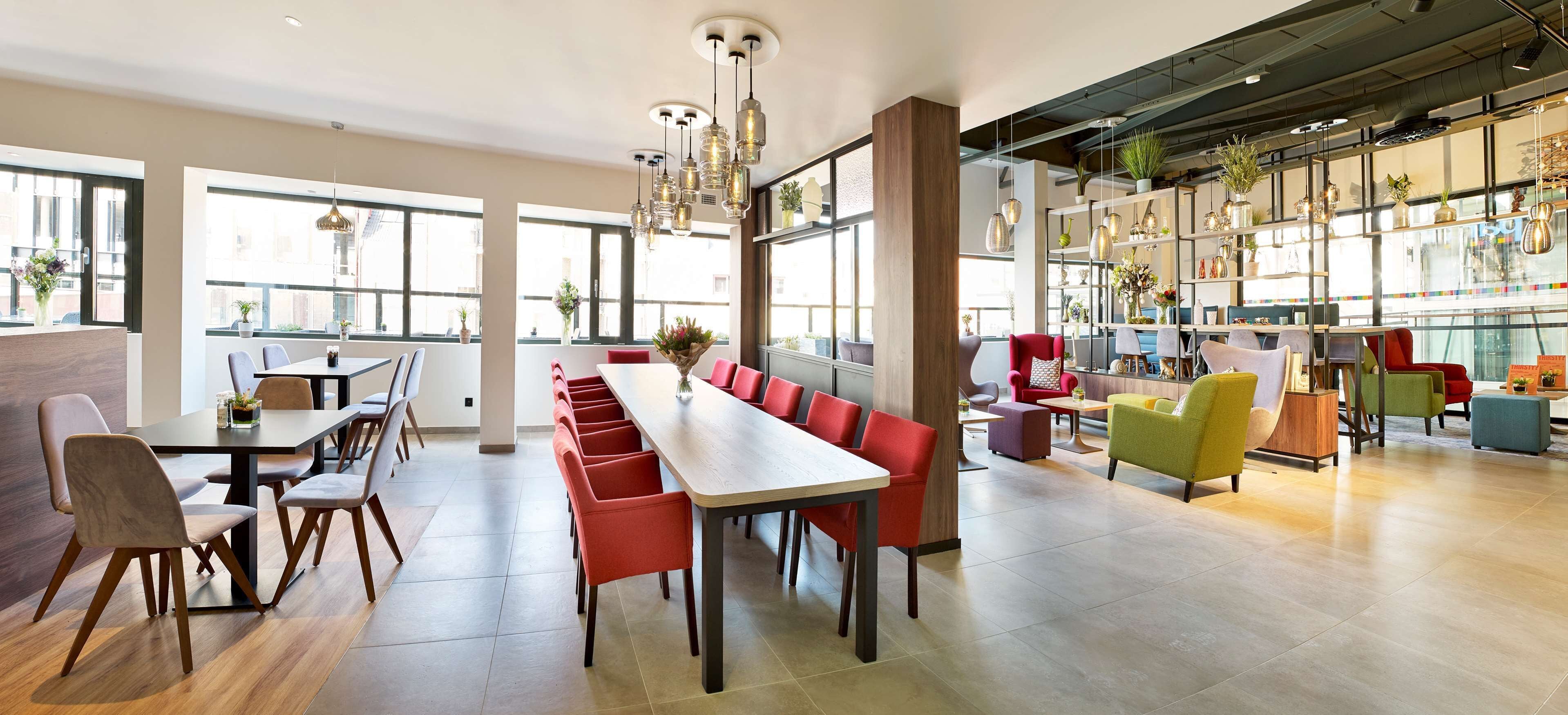 A modern dining area with long communal tables and red chairs, a bar area with colorful seating, and large windows providing natural light.