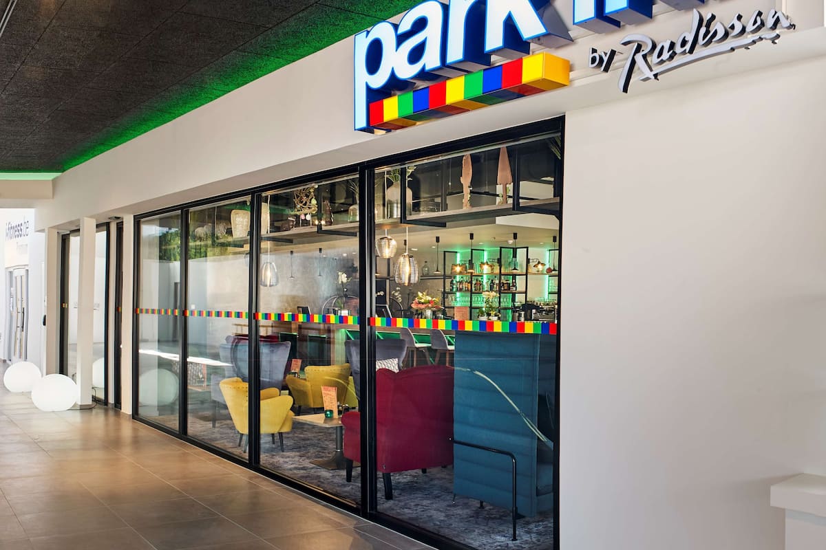 Un hotel Park Inn con diseño moderno, que destaca por un letrero iluminado grande, paredes de vidrio y diversas opciones de asientos en su interior.