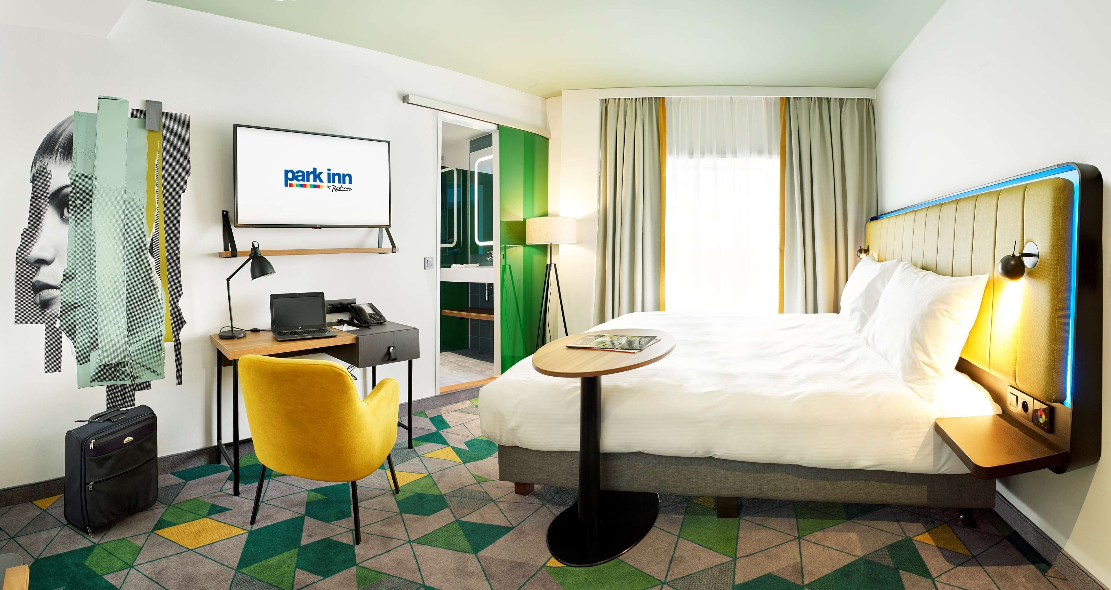 A hotel room with a bed, a desk with a computer, a chair, a lamp, and a view of the outside.